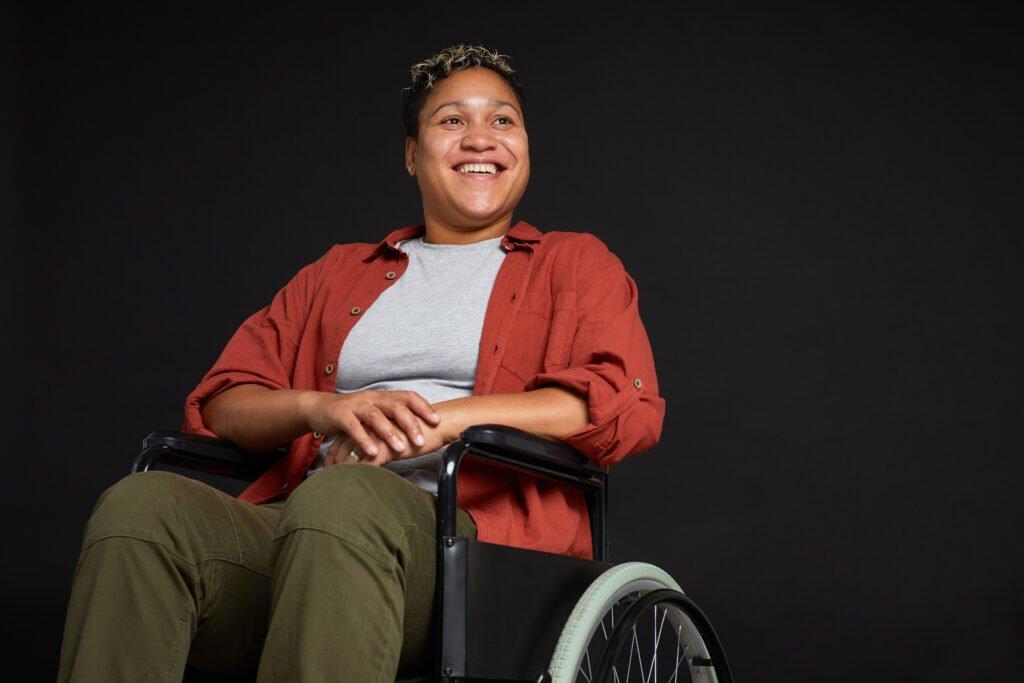 Young African woman with disabilities sitting in wheelchair and smiling isolated on black background