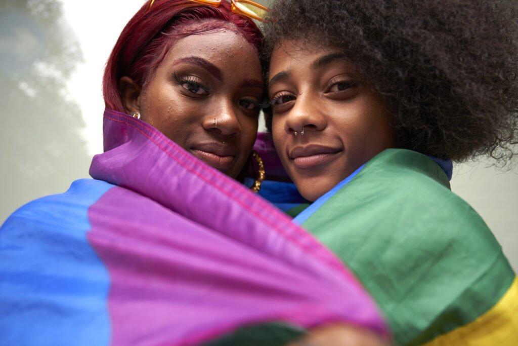 Two Black women embracing, wrapped in Pride flag.